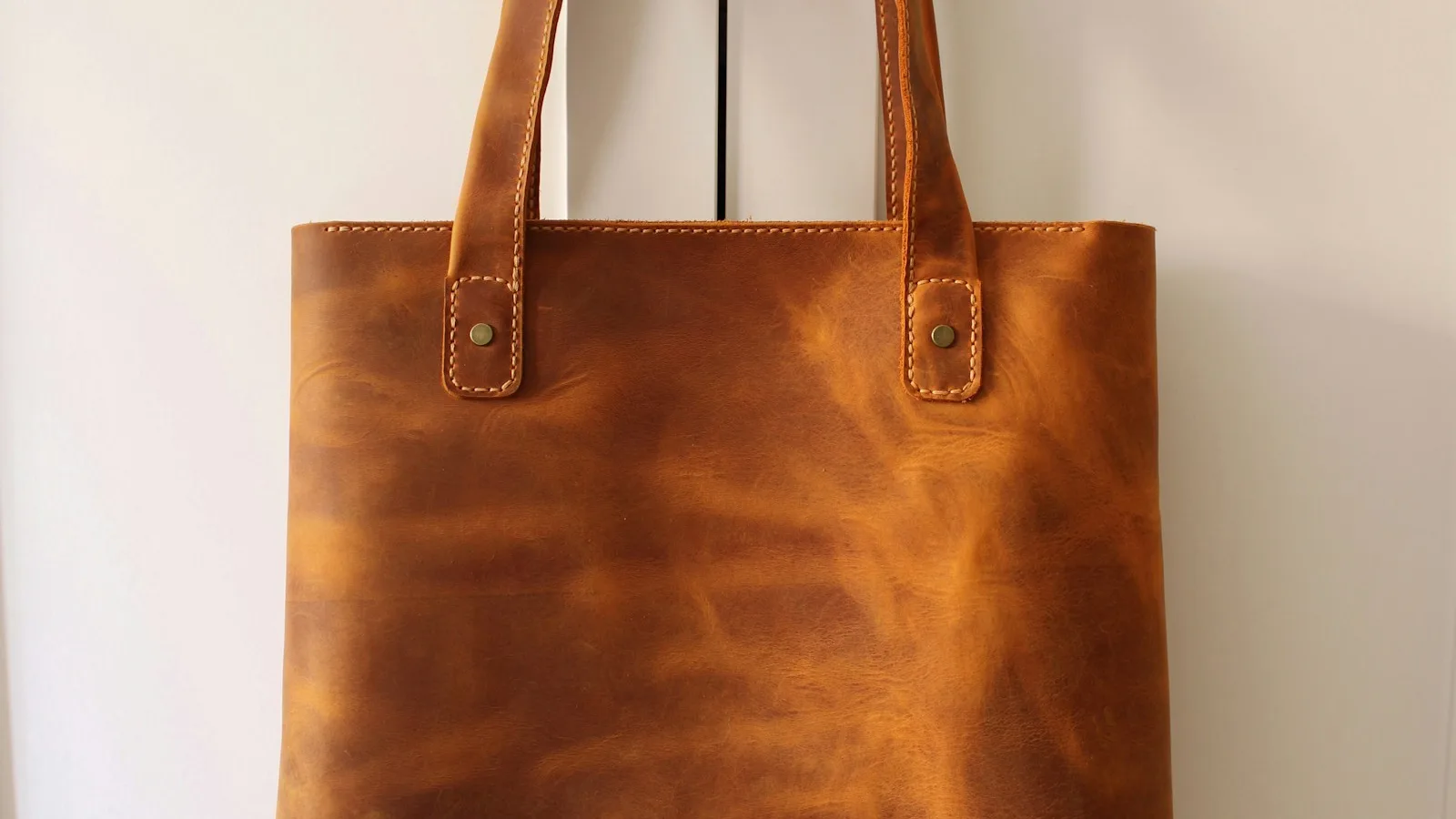 A tan leather tote bag hanging on a white door